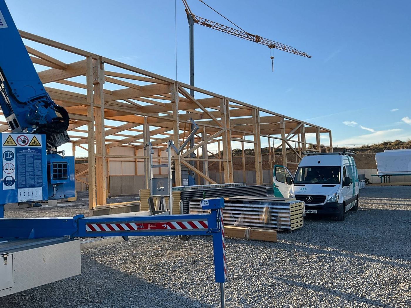 Baustelle mit Turmdrehkran und aufgerichtetem Holztragwerk einer Halle, davor ein Transporter von Holzbau Kappler.