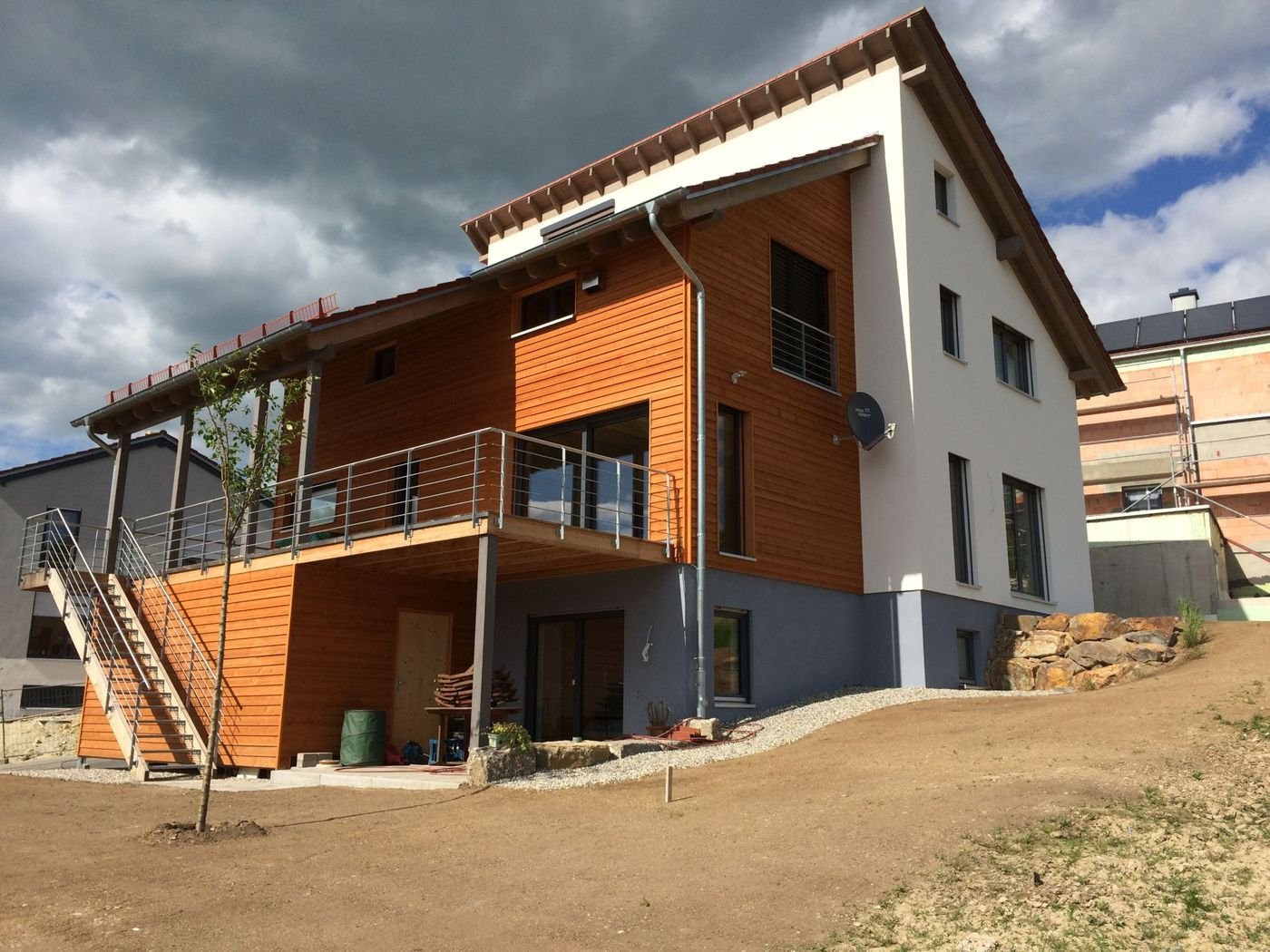 Fertiggestelltes Einfamilienhaus am Hang mit Holzfassade, weißem Putz, Satteldach und überdachter Terrasse.